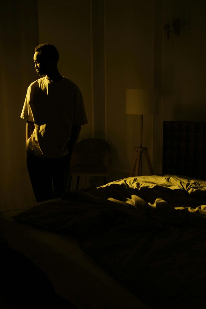 A man stands thoughtfully in a dark bedroom, illuminated by soft natural light.